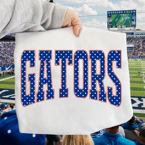 Puede incluir: Una camiseta blanca con la palabra GATORS en letras azules grandes y negritas con un patrón de puntos blancos. El texto está delineado con un borde naranja fino y centrado en la parte delantera de la prenda, que se sostiene frente a un estadio.