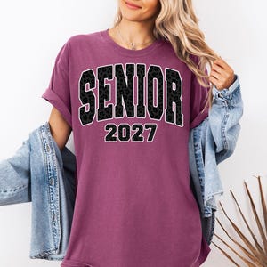 May include: A plum-colored short-sleeve t-shirt featuring the word SENIOR in a black leopard print font with a white outline, followed by the year 2027 in a matching black leopard print font with a white outline, layered under a blue denim jacket.