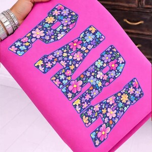 Op de afbeelding: Een felroze stof met het woord "PARIS" in grote letters. De letters zijn gevuld met een kleurrijk bloemenpatroon met roze, gele, blauwe en paarse bloemen. De stof wordt door een persoon vastgehouden.