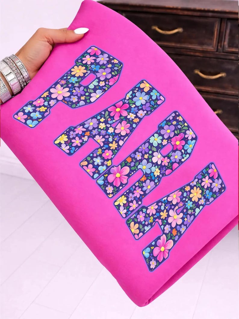 Op de afbeelding: Een felroze stof met het woord "PARIS" in grote letters. De letters zijn gevuld met een kleurrijk bloemenpatroon met roze, gele, blauwe en paarse bloemen. De stof wordt door een persoon vastgehouden.