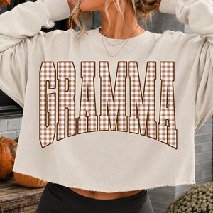 Op de afbeelding: Een beige cropped sweatshirt met lange mouwen en een rafelige zoom. Op de voorzijde staat het woord GRAMMA in grote, gebogen blokletters, gevuld met een bruin-wit vichy-ruitpatroon en omlijnd met een donkerbruine rand.
