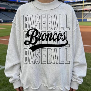 May include: A light gray sweatshirt with the word "BASEBALL" stacked above and below the word "Broncos" in a black script font. The sweatshirt has a crew neck and a slightly ruffled hemline. The background shows a baseball field.