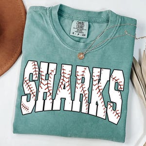 Puede incluir: Una camiseta Comfort Colors de color verde azulado con la palabra "SHARKS" en letras blancas grandes, cada una rellena con un diseño de costura de béisbol. Un collar plateado con un pequeño colgante está drapeado sobre la camisa.