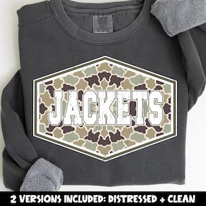 May include: A charcoal gray crewneck sweatshirt featuring a central hexagonal graphic with a camouflage pattern in shades of green, brown, and tan. The word JACKETS is printed in large, white, block-style letters across the center of the hexagon.