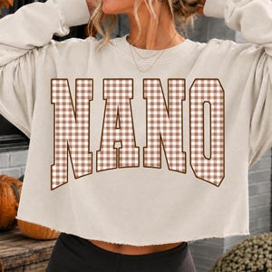 May include: A beige cropped sweatshirt featuring the word NANO in large, block-style letters. The letters have a brown and white gingham check pattern with a dark brown outline. The garment has long sleeves and a raw, unfinished hemline.