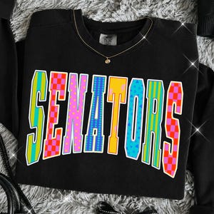 May include: A black crewneck sweatshirt featuring the word SENATORS in large, arched, multi-colored block letters. Each letter displays a unique pattern, including checkered, polka dot, striped, and geometric designs in bright neon colors.