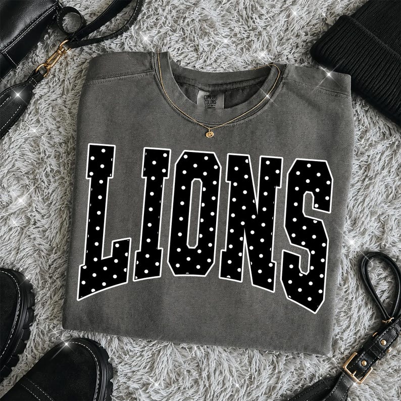 May include: A charcoal gray crewneck t-shirt featuring the word LIONS in large, bold, black varsity-style letters with a white polka dot pattern. The shirt has a ribbed neckline and a Comfort Colors brand label visible at the inner collar.