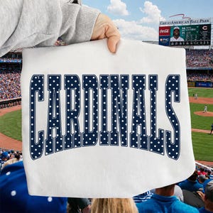 Może przedstawiać: Biały t-shirt z napisem CARDINALS wykonanym dużymi, ciemnoniebieskimi literami w białe kropki. Tekst jest wygięty w łuk na środku odzieży, kt&oacute;ra jest trzymana na tle profesjonalnego stadionu baseballowego.