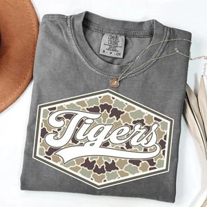 Könnte beinhalten: Graues Comfort Colors T-Shirt mit weißem und Camouflage-Design mit der Aufschrift "Tigers". Eine silberne Halskette mit einem kleinen Anhänger liegt auf dem Shirt. Das Shirt ist auf einer weißen Oberfläche gefaltet.