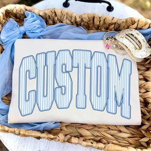 May include: Cream-colored t-shirt with the word "CUSTOM" in large, blue and white striped letters. The shirt is inside a woven basket with a blue ribbon and jewelry.