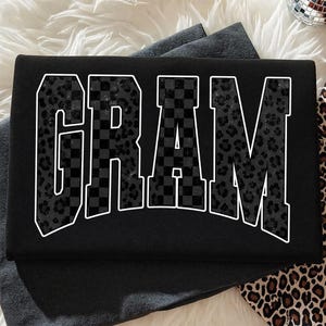 May include: Black t-shirt with the word "GRAM" in a white outline. The letters are filled with a mix of checkered and leopard print patterns. The shirt is laying on a white and gray surface.