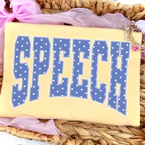 May include: A pale yellow t-shirt featuring the word SPEECH in large, blue, varsity-style block letters with a white polka dot pattern. The letters are arranged in a slight arch across the center of the shirt, which is folded neatly.