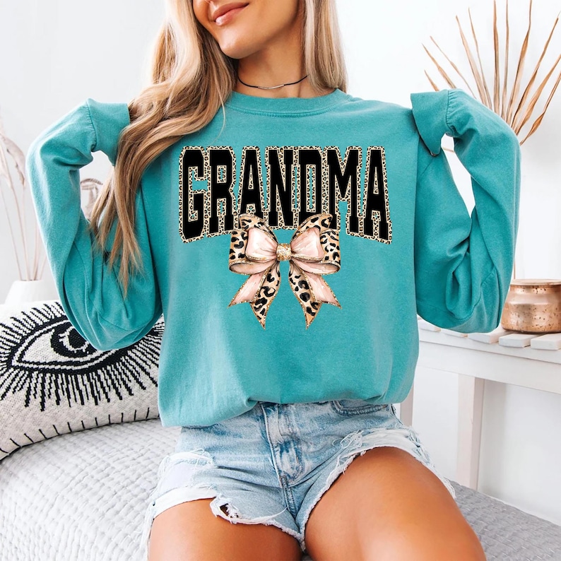 May include: A teal sweatshirt with the word "GRANDMA" in black block letters with a leopard print fill. A bow with a leopard print center and pink ribbon accents is below the word. The sweatshirt has long sleeves.