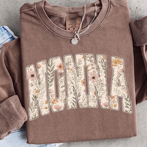 May include: A brown sweatshirt with the word "NONNA" in large, floral-patterned letters across the front. The letters are filled with a design of pink, yellow, and green flowers and leaves. A silver necklace with a pendant is visible.