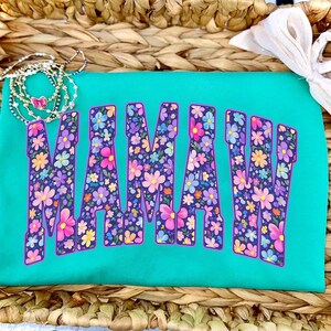 Puede incluir: Una camiseta turquesa con la palabra "MAMA" en letras grandes llenas de flores. Las letras est&aacute;n delineadas en morado y llenas de una variedad de flores coloridas. Un delicado collar y un lazo blanco est&aacute;n en el fondo.