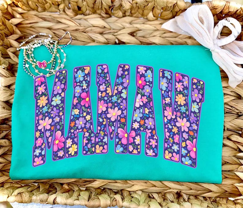 Puede incluir: Una camiseta turquesa con la palabra "MAMA" en letras grandes llenas de flores. Las letras est&aacute;n delineadas en morado y llenas de una variedad de flores coloridas. Un delicado collar y un lazo blanco est&aacute;n en el fondo.