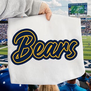 May include: White fabric with the word "Bears" in navy blue script with a gold outline. The image is held up in front of a football stadium with a scoreboard that says "Kroger Field".