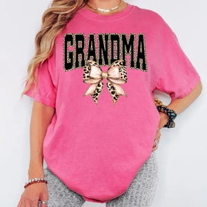 May include: A bright pink t-shirt with the word "GRANDMA" in black block letters with a leopard print pattern. A bow with a leopard print pattern is centered below the word.