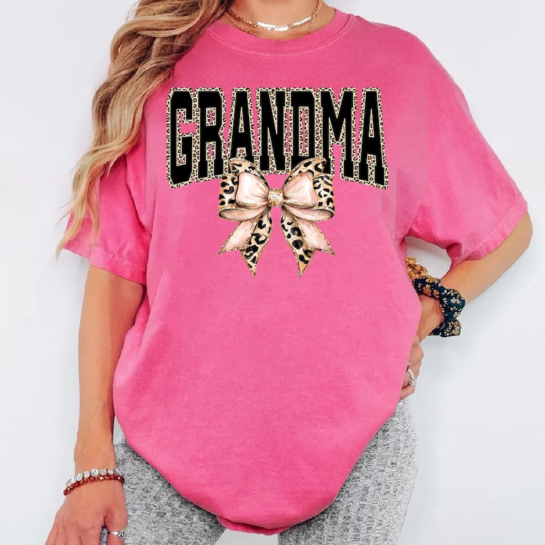 May include: A bright pink t-shirt with the word "GRANDMA" in black block letters with a leopard print pattern. A bow with a leopard print pattern is centered below the word.