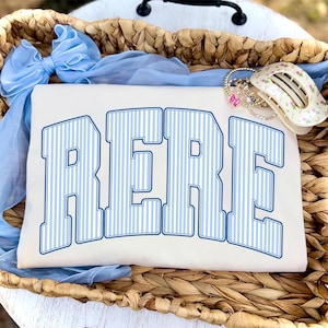 Puede incluir: Camiseta color crema con la palabra "RERE" en letras grandes a rayas azules. La camiseta está dentro de una cesta tejida, con un lazo azul y pulseras. Las letras tienen un estilo universitario.