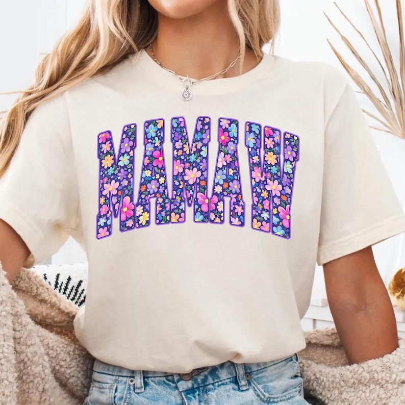 Puede incluir: Camiseta color crema con la palabra "MAMAW" en letras grandes y florales. Las letras est&aacute;n delineadas en morado oscuro, y las flores son una mezcla de rosa, azul, amarillo y morado. La camiseta tiene mangas cortas.