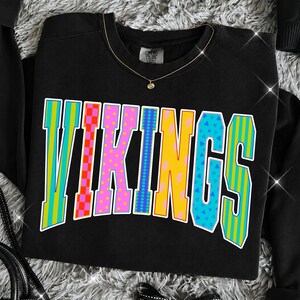 May include: A black crewneck sweatshirt featuring the word VIKINGS in large, multi-colored block letters. Each letter displays a unique pattern, including vertical stripes, red and orange checkers, pink polka dots, blue dots, yellow lightning bolts, and blue triangles.