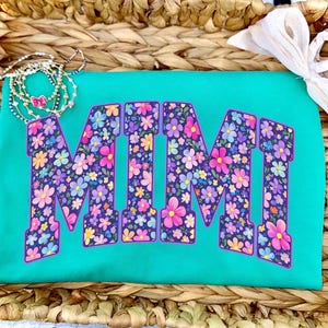May include: A turquoise t-shirt with the word "MIMI" in large, floral-patterned letters. The letters are filled with pink, purple, and yellow flowers. A delicate necklace with a butterfly charm and a white bow are also visible.