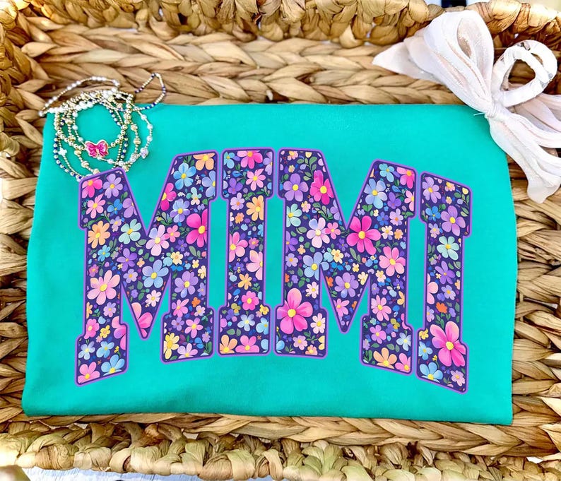 May include: A turquoise t-shirt with the word "MIMI" in large, floral-patterned letters. The letters are filled with pink, purple, and yellow flowers. A delicate necklace with a butterfly charm and a white bow are also visible.