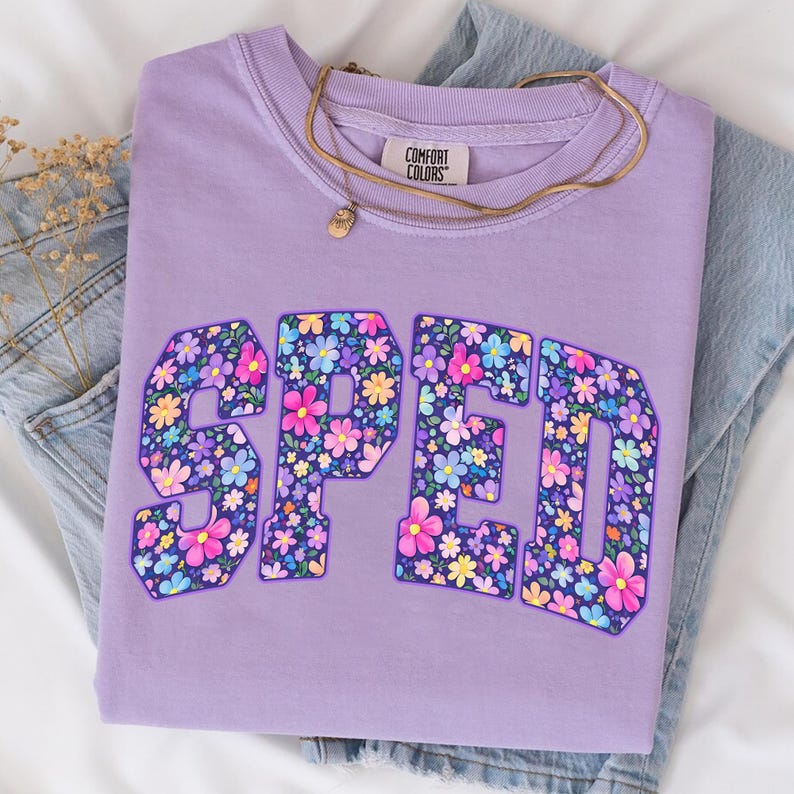 Pode incluir: Uma camisola lil&aacute;s Comfort Colors com a palavra "SPED" num design floral. Um colar de ouro com um pequeno pendente repousa sobre a camisa. A camisola est&aacute; dobrada sobre um par de cal&ccedil;as de ganga azuis.