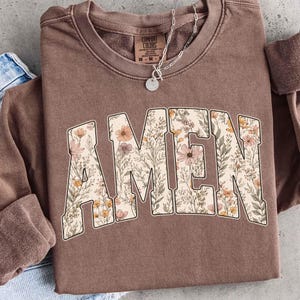 May include: A brown Comfort Colors sweatshirt with the word "AMEN" in large, floral-filled letters. The letters are filled with a variety of flowers in shades of pink, orange, and cream. A silver necklace with a pendant is visible.