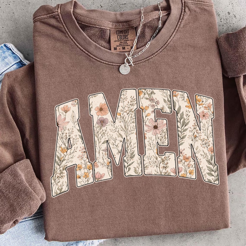May include: A brown Comfort Colors sweatshirt with the word "AMEN" in large, floral-filled letters. The letters are filled with a variety of flowers in shades of pink, orange, and cream. A silver necklace with a pendant is visible.