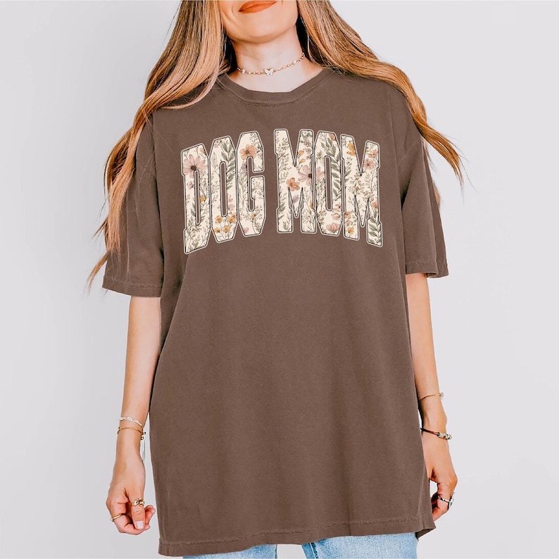 Op de afbeelding: Bruin t-shirt met de tekst "DOG MOM" in een bloemenontwerp. De tekst is over de borst gebogen. Het shirt heeft korte mouwen en een relaxte pasvorm.