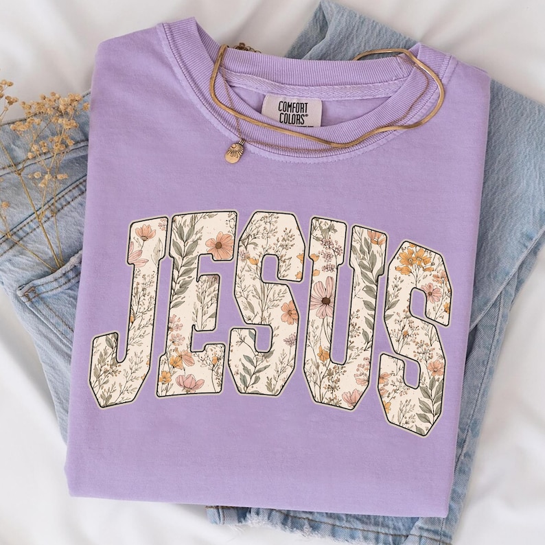 Peut inclure: Un sweat-shirt lilas avec le mot "JESUS" dans un motif floral. Les lettres sont remplies d'un motif de fleurs et de feuilles dans des tons p&ecirc;che, cr&egrave;me et vert. Un collier dor&eacute; repose sur le t-shirt, pli&eacute; sur un jean bleu.