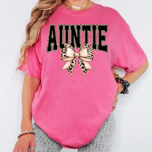 May include: Auntie graphic tee in a vibrant pink hue. The word "AUNTIE" is printed across the front in black letters with a leopard print pattern. A bow with a leopard print design is centered below the word.