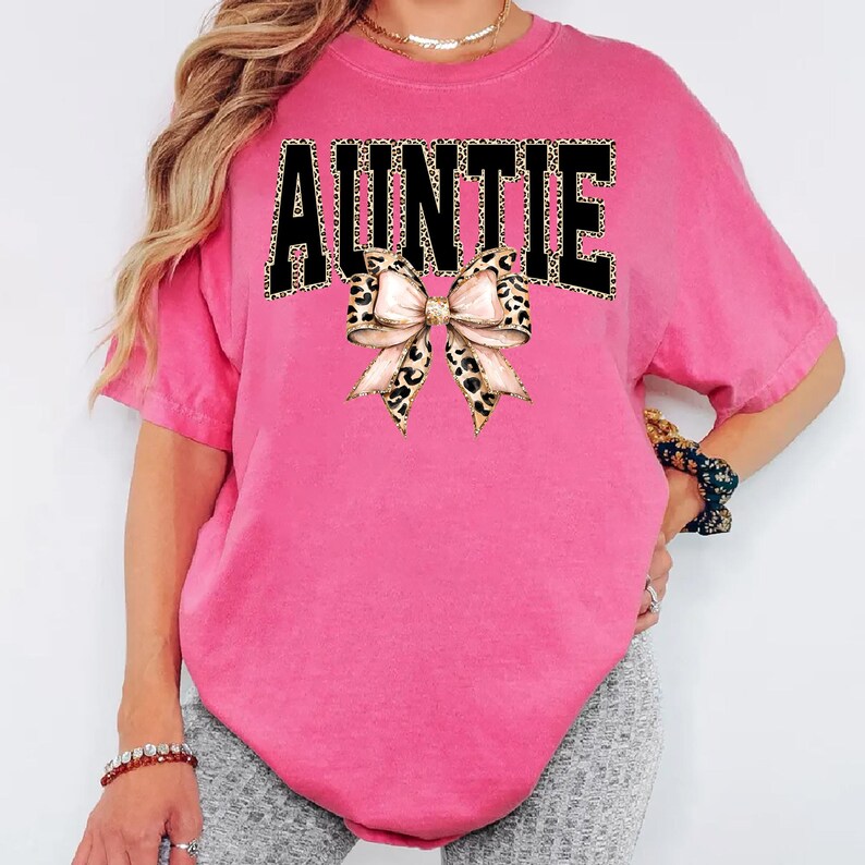 May include: Auntie graphic tee in a vibrant pink hue. The word "AUNTIE" is printed across the front in black letters with a leopard print pattern. A bow with a leopard print design is centered below the word.