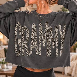 Pode incluir: Uma camisola de manga comprida em cinzento-carvão com uma bainha curta de corte cru. A parte da frente apresenta a palavra GRAMMY impressa numa fonte grande e arrojada, preenchida com um padrão de leopardo em bege e preto.