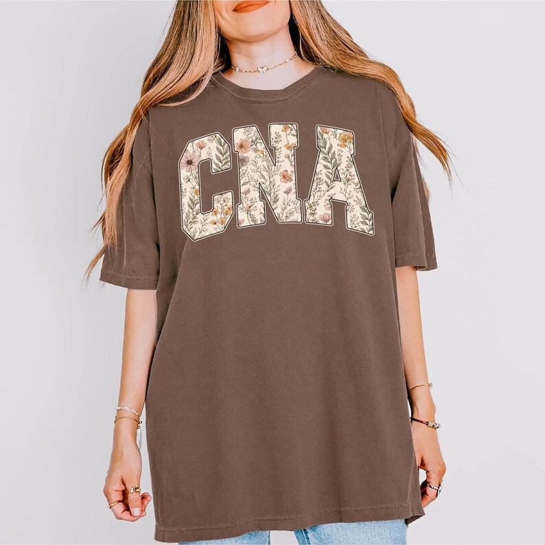 May include: A brown t-shirt with the letters "CNA" in a floral design. The letters are filled with a pattern of flowers and leaves in shades of pink, yellow, and green. The shirt has short sleeves and a relaxed fit.