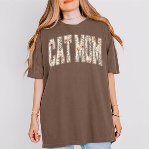 May include: A brown t-shirt featuring the text "CAT MOM" in a floral design. The shirt has short sleeves and a relaxed fit. The text is in a white, arched font with a floral pattern.