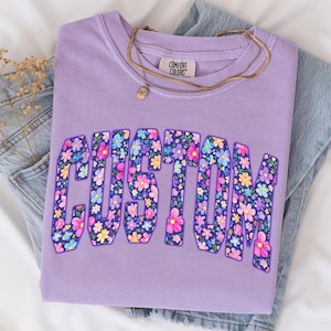 May include: A lilac Comfort Colors sweatshirt with the word "CUSTOM" in a floral design. The floral design features a variety of colorful flowers. A gold necklace with a pendant is draped across the shirt, which is folded on top of blue denim.