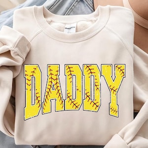 Op de afbeelding: Crèmekleurige sweatshirt met het woord "DADDY" in grote, gele, verweerde letters. De letters zijn gevuld met een honkbalsteekpatroon en zwart omlijnd. Een casual, comfortabele top.