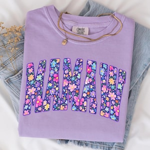 Puede incluir: Una sudadera Comfort Colors lavanda con la palabra "MAMA" en un dise&ntilde;o floral. Un collar dorado con un peque&ntilde;o colgante descansa sobre la camisa. La camisa est&aacute; doblada sobre un par de jeans azules.