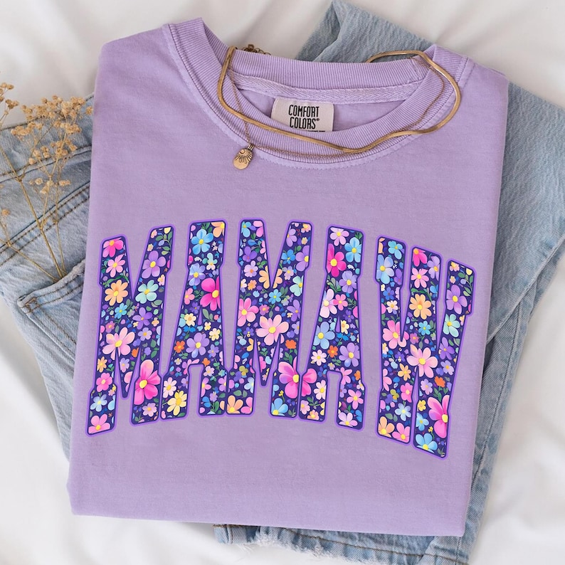 Puede incluir: Una sudadera Comfort Colors lavanda con la palabra "MAMA" en un dise&ntilde;o floral. Un collar dorado con un peque&ntilde;o colgante descansa sobre la camisa. La camisa est&aacute; doblada sobre un par de jeans azules.