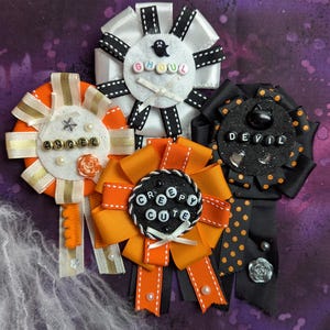 Può includere: Quattro rosette a nastro circolari con temi di Halloween. Una rosetta bianca riporta la scritta GHOUL, una arancione e oro ANGEL, una arancione CREEPY CUTE e una nera a pois arancioni DEVIL. Ognuna include perline, ciondoli e nastri in tessuto.