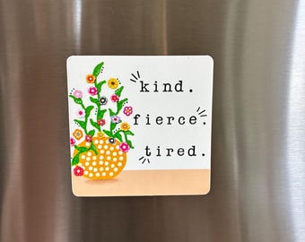 May include: A square magnet featuring a white background with a yellow polka-dot vase containing green stems and colorful flowers. The text reads: kind. fierce. tired. The design includes small black accent marks around the words and flowers.