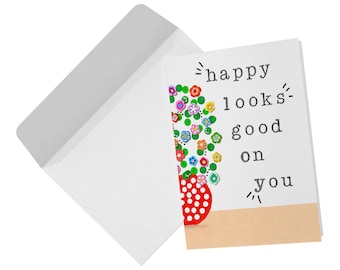 May include: A white greeting card and matching envelope. The card features a red vase with white polka dots containing a bouquet of colorful small flowers. The text "happy looks good on you" is printed in black typewriter-style font on the right.