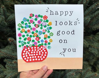 May include: A square greeting card featuring a red, white-polka-dotted vase filled with green dots and colorful floral embellishments. The text "happy looks good on you" is printed in black typewriter-style font on the right side of the card.
