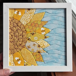 May include: A framed fabric art piece featuring a sunflower design. The sunflower has yellow petals with bee and floral patterns, a brown center, and a blue wavy background. The frame is white.
