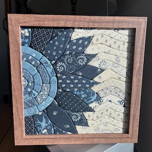 May include: A framed textile art piece featuring a stylized sunflower design. The artwork uses various blue and cream-colored patterned fabrics, creating a textured, layered effect. The frame is made of wood.