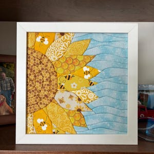 May include: A framed fabric art piece featuring a half sunflower design. The sunflower has yellow and brown petals with bee and honeycomb patterns. The other half is light blue with wavy lines. The artwork is in a white frame.