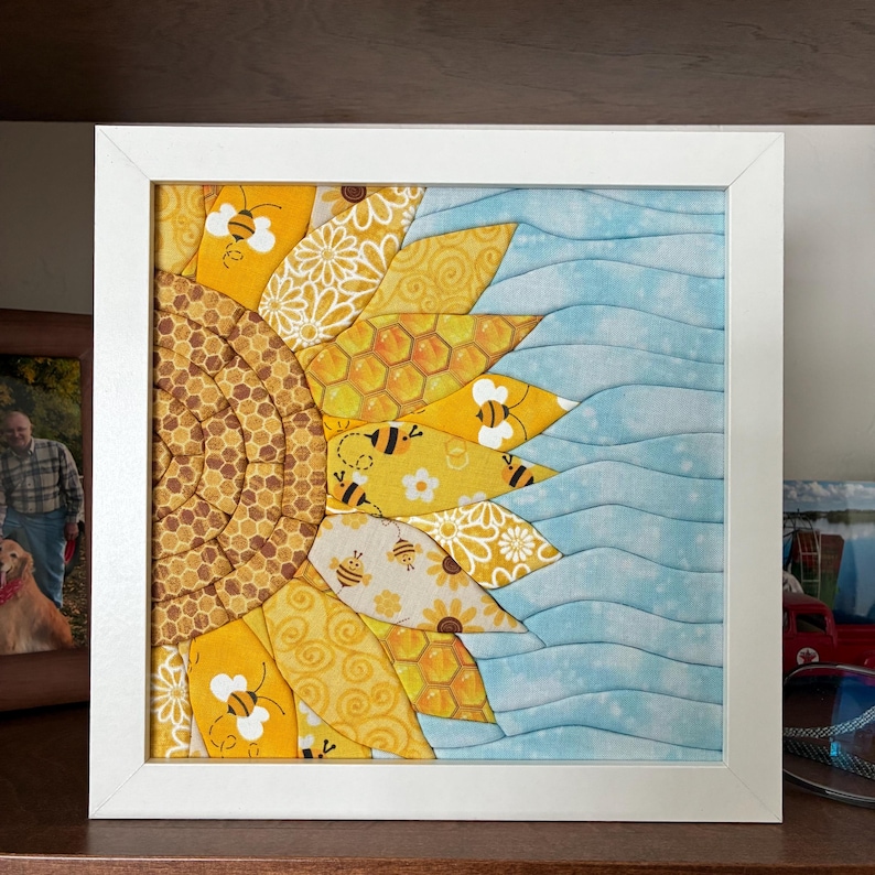 May include: A framed fabric art piece featuring a half sunflower design. The sunflower has yellow and brown petals with bee and honeycomb patterns. The other half is light blue with wavy lines. The artwork is in a white frame.